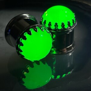 May include: Two green glow-in-the-dark ear gauges with silver metal accents. The gauges have a crown design around the green glass.