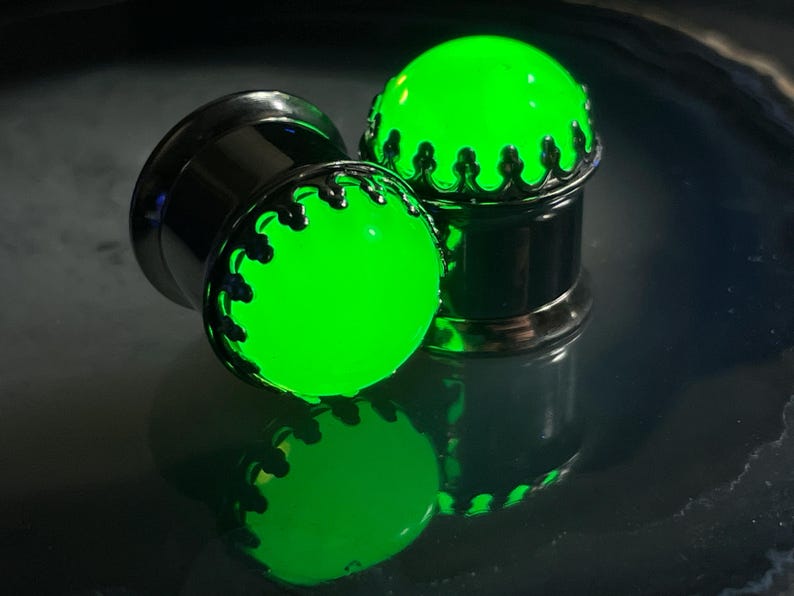 May include: Two green glow-in-the-dark ear gauges with silver metal accents. The gauges have a crown design around the green glass.