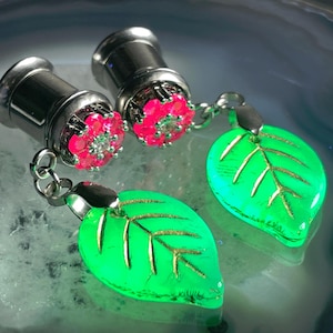 Puede incluir: Dos expansores de oreja plateados con detalles florales rosas brillantes y colgantes de hojas verdes. Las hojas tienen detalles de vetas doradas. Expansores para piercings de oreja estirados.