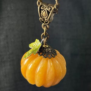 May include: A pendant necklace featuring a small, orange pumpkin charm. The pumpkin has a ribbed texture and is topped with a green leaf and a decorative bronze-colored cap. The chain and clasp are also bronze-colored.