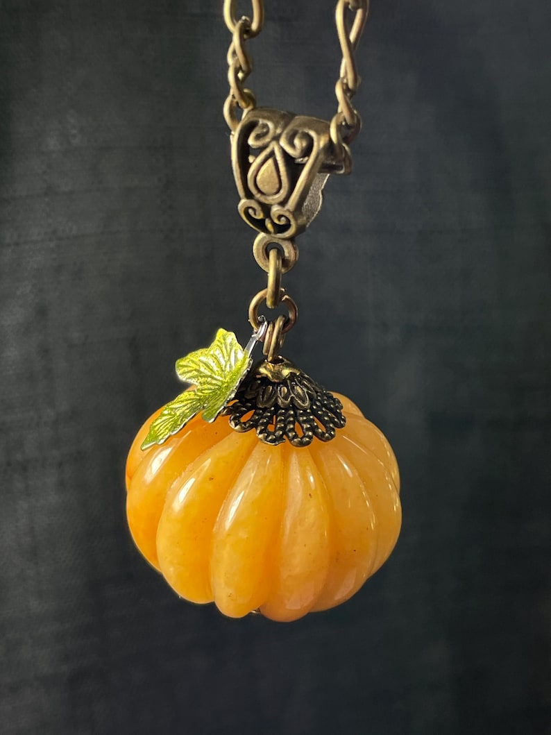 May include: A pendant necklace featuring a small, orange pumpkin charm. The pumpkin has a ribbed texture and is topped with a green leaf and a decorative bronze-colored cap. The chain and clasp are also bronze-colored.