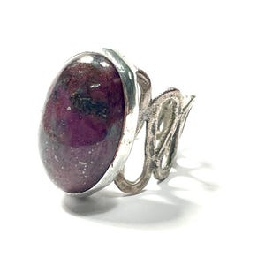 May include: A silver ring with a large oval-shaped gemstone. The gemstone is a deep purple color with flecks of silver and black.