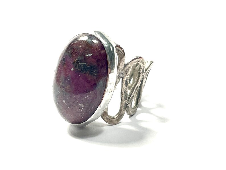 May include: A silver ring with a large oval-shaped gemstone. The gemstone is a deep purple color with flecks of silver and black.