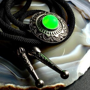 May include: A bolo tie featuring a silver-toned, ornate pendant with a vibrant green center stone. The pendant is connected to a black braided cord. Two decorative silver-toned tips complete the design, creating a classic Western accessory.