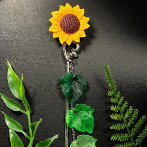 May include: A sunflower-themed accessory with a bright yellow flower and a dark brown center. The design includes green leaf accents and a silver-colored metal stem. The background is a dark, textured surface.