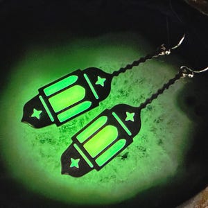 May include: A pair of black lantern earrings with a glowing green interior and star accents. The earrings have a long, textured chain and a silver hook. The background is a glowing green.