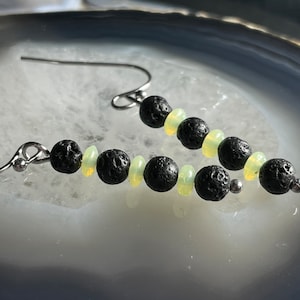 Puede incluir: Pendientes colgantes con cuentas de roca volcánica negra y pequeñas cuentas verde claro, ensartadas en ganchos plateados. Diseño sencillo y natural.