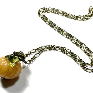 May include: A pumpkin-shaped pendant necklace with a bronze-colored chain. The pumpkin is a light orange color with green and gold leaf accents. The chain is a linked style, and the necklace is displayed on a white background.