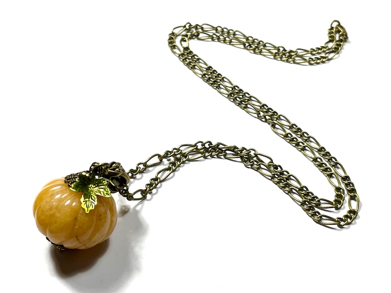 May include: A pumpkin-shaped pendant necklace with a bronze-colored chain. The pumpkin is a light orange color with green and gold leaf accents. The chain is a linked style, and the necklace is displayed on a white background.