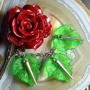 May include: A dark red rose-shaped brooch with a silver stem and chain, connected to three green leaf charms, each with a silver spike. The brooch is set against a marbled background.