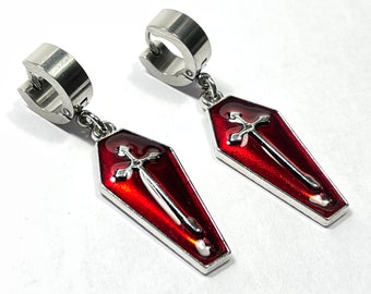 May include: A pair of silver-tone earrings featuring a coffin-shaped pendant with a red enamel finish. Each pendant has a silver cross and sword design. The earrings have a hinged hoop closure.