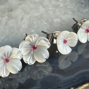 May include: A pair of white mother-of-pearl flower earrings with pink gemstone centres. The earrings are made of stainless steel and have a post back.