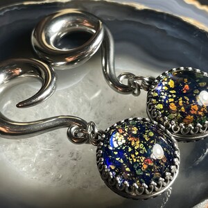 May include: A pair of silver-toned ear gauges with a spiral design. Each gauge has a dangling, round charm with a dark blue, speckled glass-like appearance, framed by a decorative silver border. The charms have gold and green flecks.