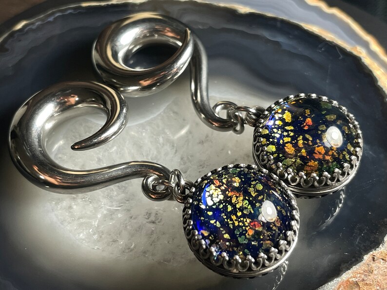 May include: A pair of silver-toned ear gauges with a spiral design. Each gauge has a dangling, round charm with a dark blue, speckled glass-like appearance, framed by a decorative silver border. The charms have gold and green flecks.