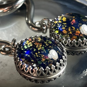 May include: Close-up of two silver pendants with a dark blue, speckled glass cabochon. The glass has gold, green, and orange flecks. The pendants have a decorative silver crown setting and are attached to a silver chain.