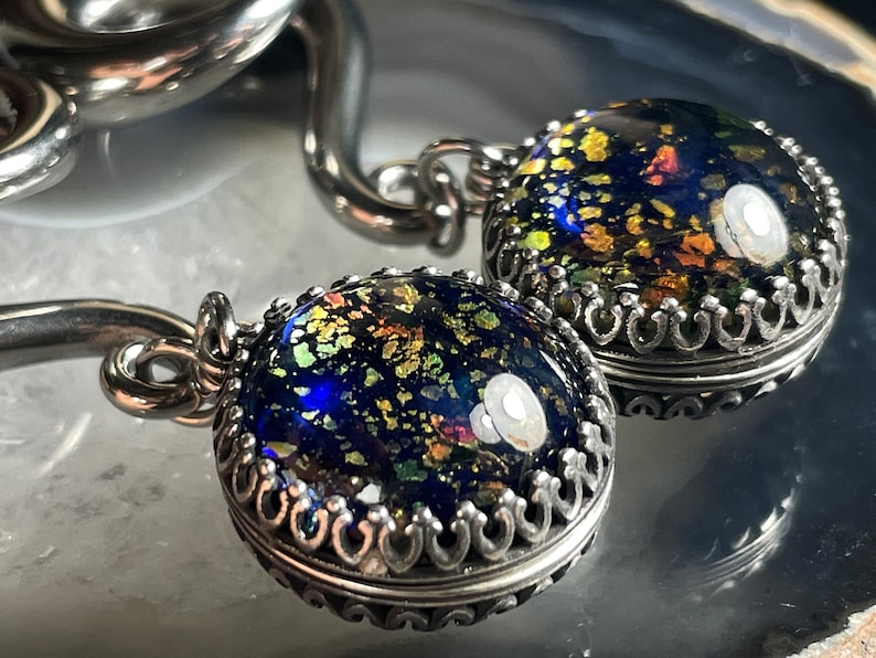 May include: Close-up of two silver pendants with a dark blue, speckled glass cabochon. The glass has gold, green, and orange flecks. The pendants have a decorative silver crown setting and are attached to a silver chain.