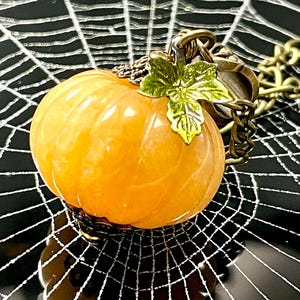 May include: A pumpkin-shaped pendant necklace with a gold-colored chain. The orange pumpkin has a green leaf accent and is set against a black background with a white spiderweb design. A festive Halloween accessory.