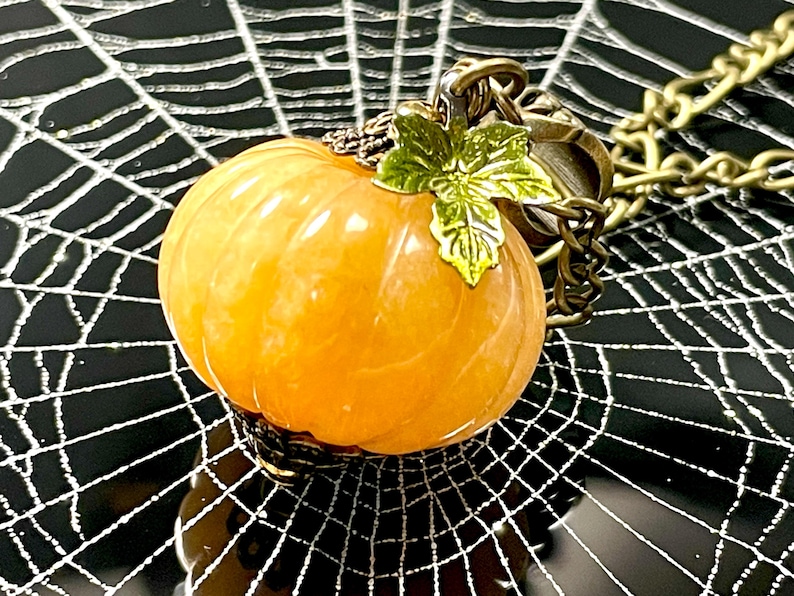 May include: A pumpkin-shaped pendant necklace with a gold-colored chain. The orange pumpkin has a green leaf accent and is set against a black background with a white spiderweb design. A festive Halloween accessory.