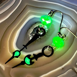 May include: A pair of unique earrings featuring black clock hands with glowing green accents and faceted green beads. The earrings have gold-colored ear wires and are set against a neutral, patterned background.
