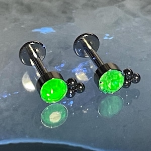 May include: Two silver body jewelry pieces with green gem stones and black beads. The jewelry is designed for ear piercings.