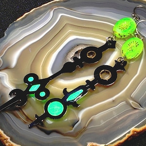May include: Two black metal earrings with clock hand designs and neon green details. The earrings have a circular design with a green centre and a green gem-like shape. The earrings are displayed on a marbled surface.
