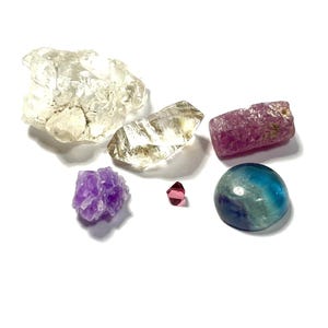 May include: A collection of raw gemstones in different shapes and colours. Includes a large clear quartz cluster, a faceted clear quartz, a pink tourmaline, a purple amethyst, a small pink crystal, and a blue and green fluorite.
