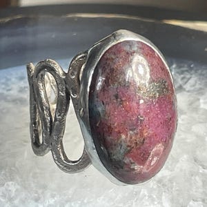 May include: A silver ring with a large, oval-shaped, pink and grey gemstone set in a bezel setting. The band of the ring is twisted and textured.