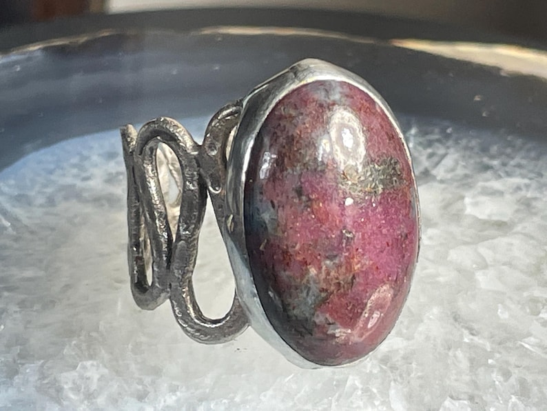 May include: A silver ring with a large, oval-shaped, pink and grey gemstone set in a bezel setting. The band of the ring is twisted and textured.