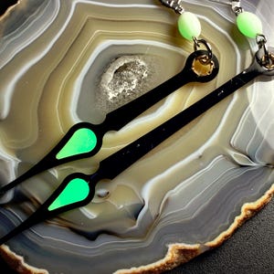 May include: Two black watch hands with a neon green accent on a layered agate stone. The hands are long and slender, with a pointed tip and a rounded base. The neon green accent is a teardrop shape.
