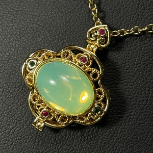 Puede incluir: Un collar con un colgante de filigrana dorada adornado con una piedra ovalada verde pálida y acentos rojos y verdes.