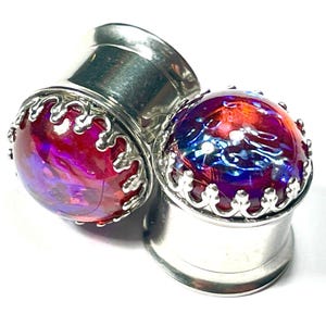 May include: A pair of silver-colored ear gauges with a vibrant, iridescent, glass-like center. The centers display a mix of red, purple, and blue hues. The edges of the gauges are adorned with a decorative, scalloped silver trim.