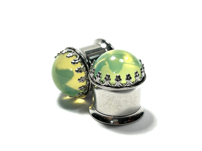 May include: A pair of green glass ear gauges with a silver crown design. The gauges are made of surgical steel and are available in a variety of sizes.