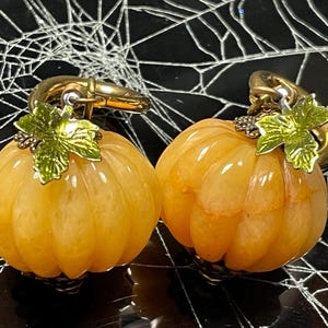 Puede incluir: Dos pendientes con forma de calabaza naranja con ganchos dorados y detalles de hojas verdes. Los pendientes se presentan sobre un fondo negro con un diseño de telaraña blanca, evocando un tema de Halloween.