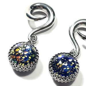 May include: Pair of silver-toned ear gauges with a spiral design. Each gauge features a round, dark blue pendant with flecks of gold, green, and yellow. The pendant is framed by a decorative silver border.