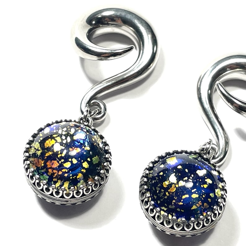 May include: Pair of silver-toned ear gauges with a spiral design. Each gauge features a round, dark blue pendant with flecks of gold, green, and yellow. The pendant is framed by a decorative silver border.