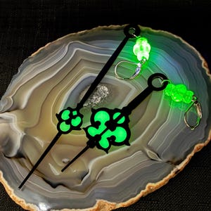 May include: A pair of black earrings with a glowing green design. The earrings have a long, slender black stem with a circular top and a decorative green center. The earrings are on a marbled surface.