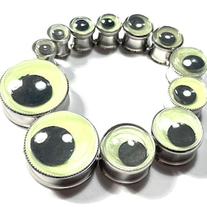 May include: A collection of silver-toned ear gauges with a unique eye design. Each gauge features a black pupil set against a pale green iris, creating a striking visual effect. The gauges vary in size, showcasing a range of options.