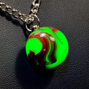 May include: A round pendant necklace with a bright green and maroon swirled design. The pendant is attached to a silver chain. The necklace is displayed on a black surface.