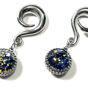 May include: A pair of silver-toned spiral earrings with dangling, round pendants. The pendants feature a dark blue background with gold and yellow flecks, framed by a decorative silver edge. The earrings are displayed against a white background.