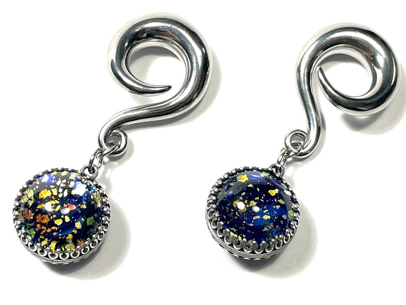 May include: A pair of silver-toned spiral earrings with dangling, round pendants. The pendants feature a dark blue background with gold and yellow flecks, framed by a decorative silver edge. The earrings are displayed against a white background.