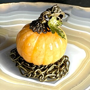 May include: A small, orange pumpkin-shaped pendant with a bronze-colored chain base and accents. The pumpkin has vertical ridges and is topped with a bronze cap and a green leaf. The pendant is a fall-themed accessory.
