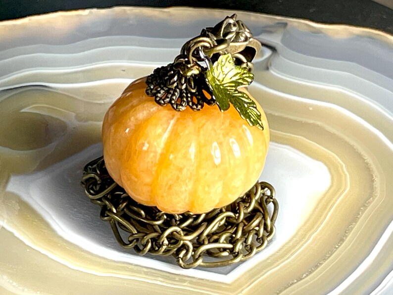May include: A small, orange pumpkin-shaped pendant with a bronze-colored chain base and accents. The pumpkin has vertical ridges and is topped with a bronze cap and a green leaf. The pendant is a fall-themed accessory.