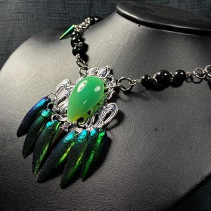 May include: A unique necklace featuring a large, teardrop-shaped green stone pendant, surrounded by ornate silver-toned metalwork. The necklace includes iridescent beetle wing accents and a black beaded chain.