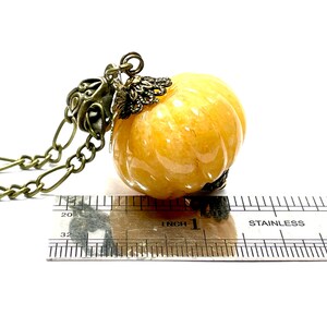 May include: A pumpkin-shaped pendant necklace with a bronze chain. The pendant is a translucent, light orange color with carved ridges. The necklace is measured with a ruler, showing the pendant is approximately 1 inch in diameter.
