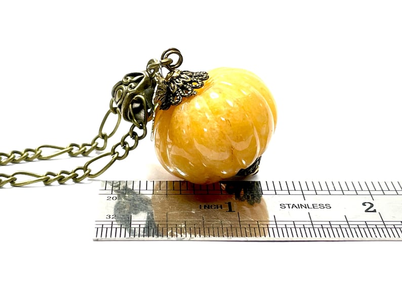 May include: A pumpkin-shaped pendant necklace with a bronze chain. The pendant is a translucent, light orange color with carved ridges. The necklace is measured with a ruler, showing the pendant is approximately 1 inch in diameter.