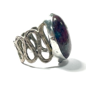 May include: A silver ring with a large, oval-shaped, dark red gemstone set in a silver bezel. The band of the ring is made of twisted silver.