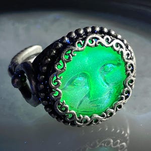 May include: A silver-toned ring with an oval, glowing green center featuring a face design. The ring has an ornate, filigree-style border and a textured band. The face within the ring is detailed, and the overall design is intricate.