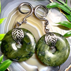 May include: A pair of ear gauges featuring polished olive green stone donuts with a mottled texture. Each gauge is attached to a silver-toned metal spiral hook and a delicate, openwork silver-toned leaf charm that hangs through the center hole.