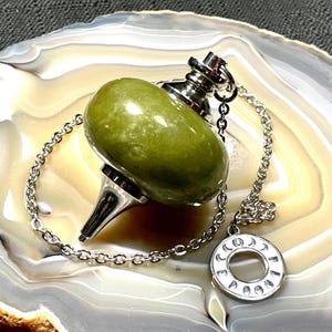 May include: A green stone pendulum with a silver chain and a circular charm. The pendulum has a rounded, oval shape and a pointed tip. The charm features a crescent moon and other symbols. The background is a polished stone surface.
