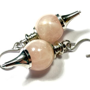 May include: A pair of rose quartz earrings. Each earring features a pale pink, spherical stone with silver-colored metal accents. The earrings have a hook closure. The background is white.